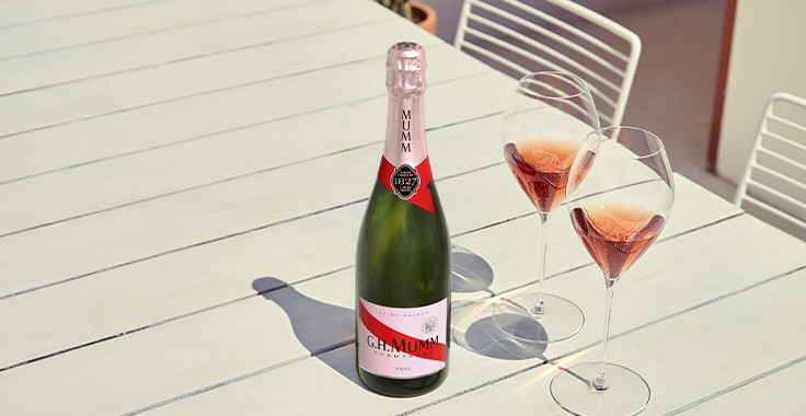 Bouteille de Champagne G.H.Mumm Rosé sur une table dans une ambiance claire d'été, avec des flûtes à champagne G.H.Mumm