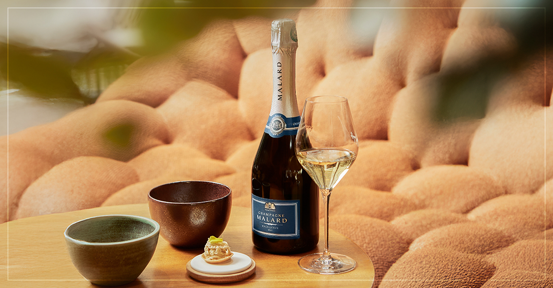 Brut Excellence Champagne Malard sur une table en bois chic dans un salon avec un canapé texturé de couleur brique